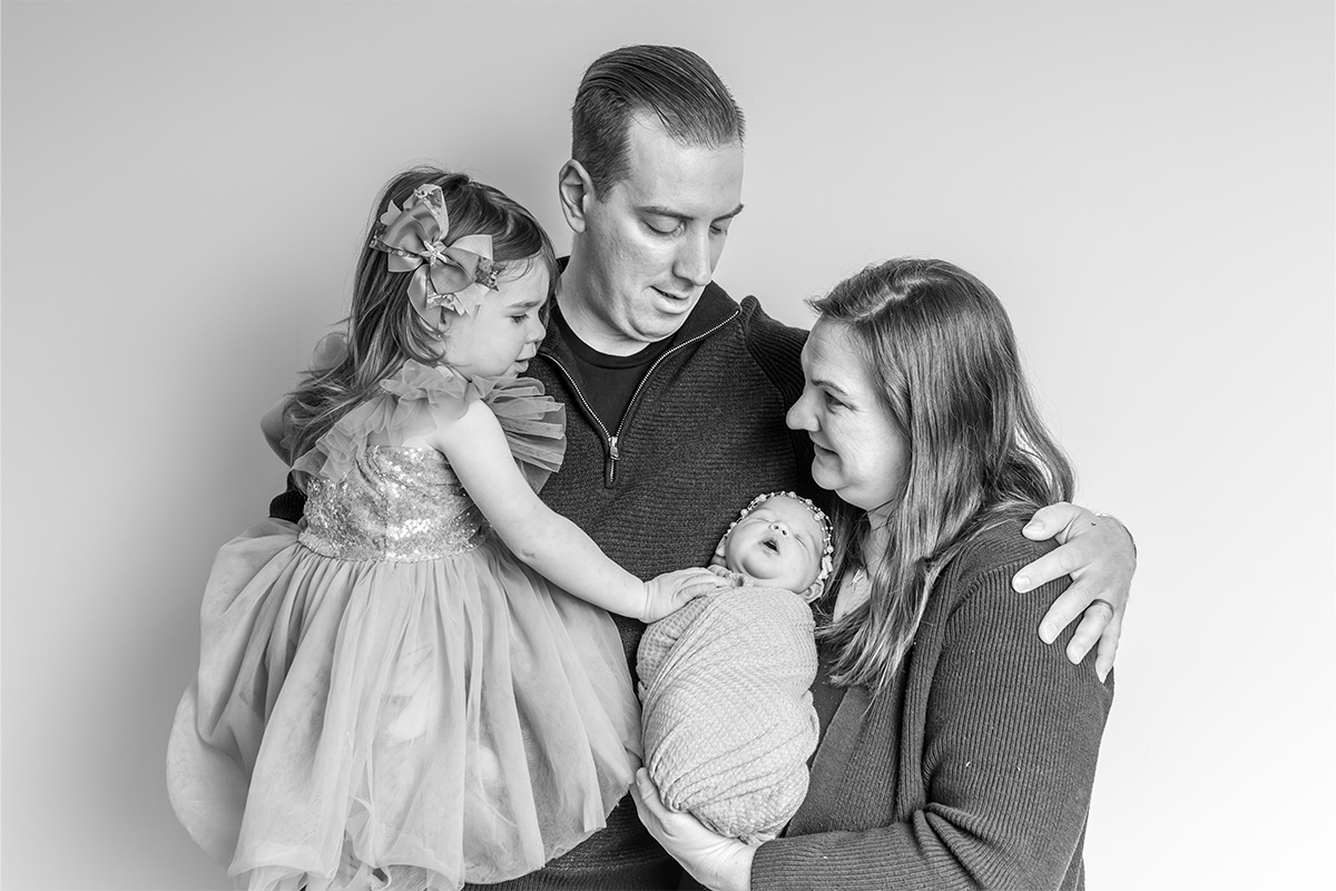 A man and woman hold a young girl in a dress and a swaddled baby, standing close together and looking at the baby—beautifully captured by a Long Island photographer.