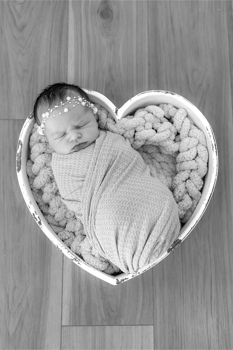 Newborn baby wrapped in a blanket, sleeping peacefully in a heart-shaped basket on a wooden floor—captured by a Long Island photographer.