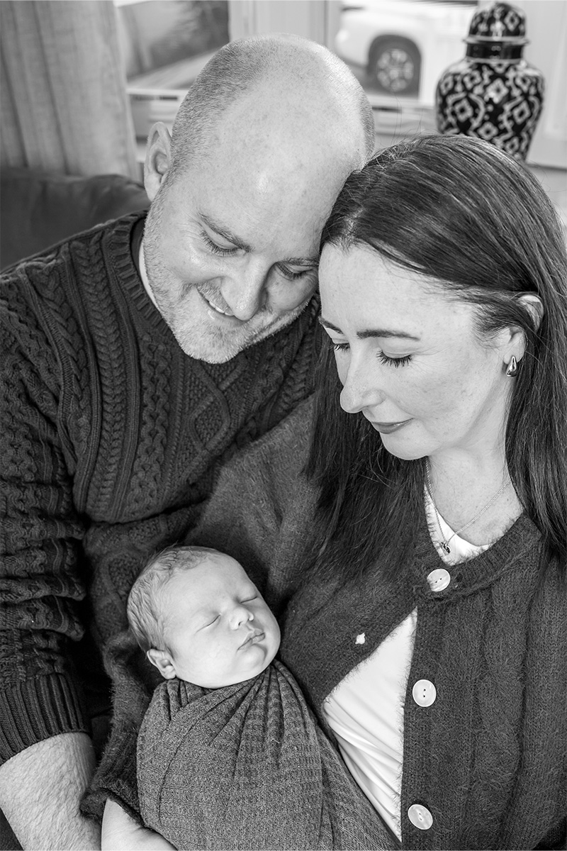 A man and woman sit closely together, looking down at a sleeping newborn wrapped in a blanket in the woman's arms, beautifully captured by a Long Island photographer.