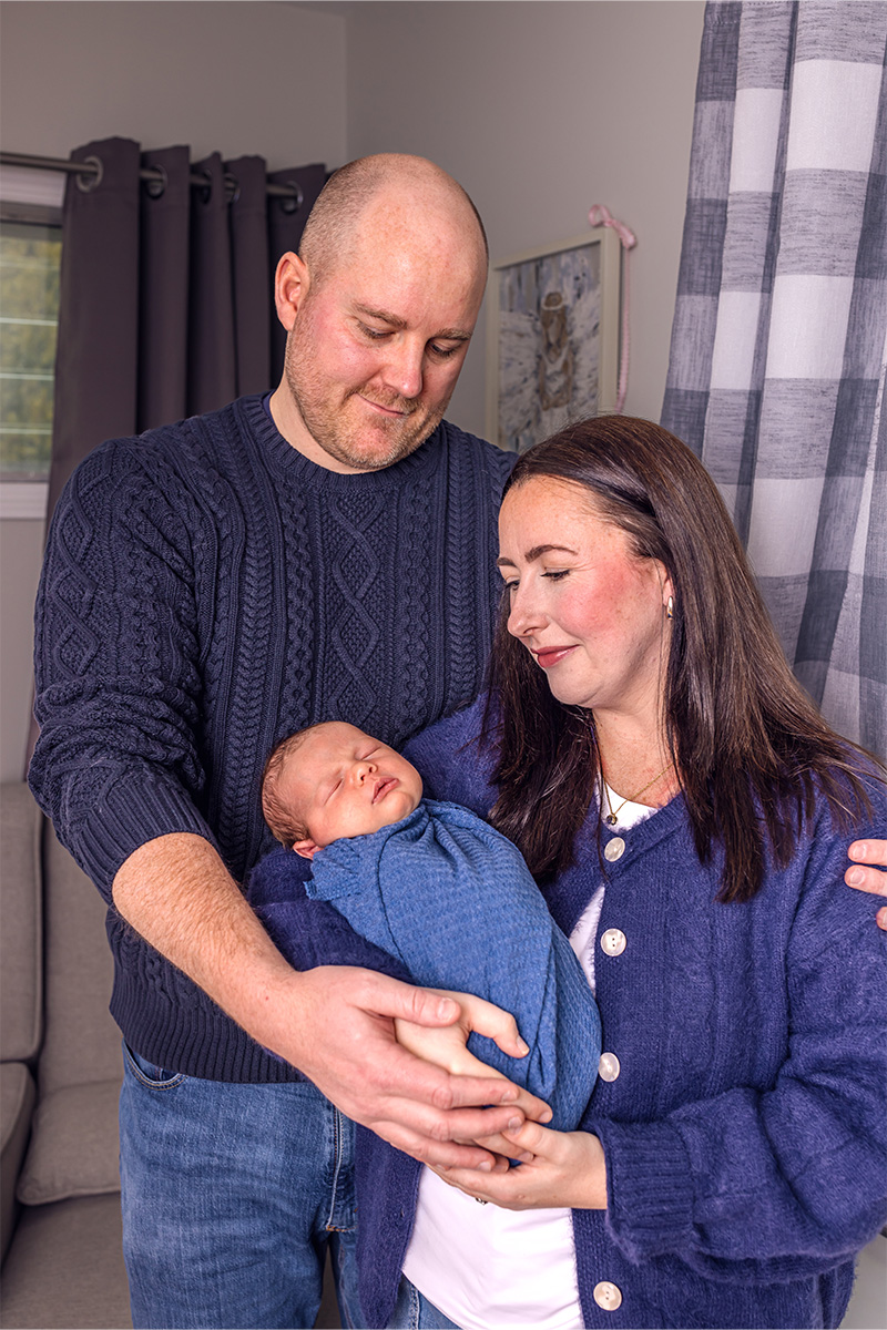 A man and woman stand close together indoors, gently holding a newborn wrapped in a blue blanket. All three are wearing blue tones and looking at the baby, beautifully captured by a Long Island photographer.