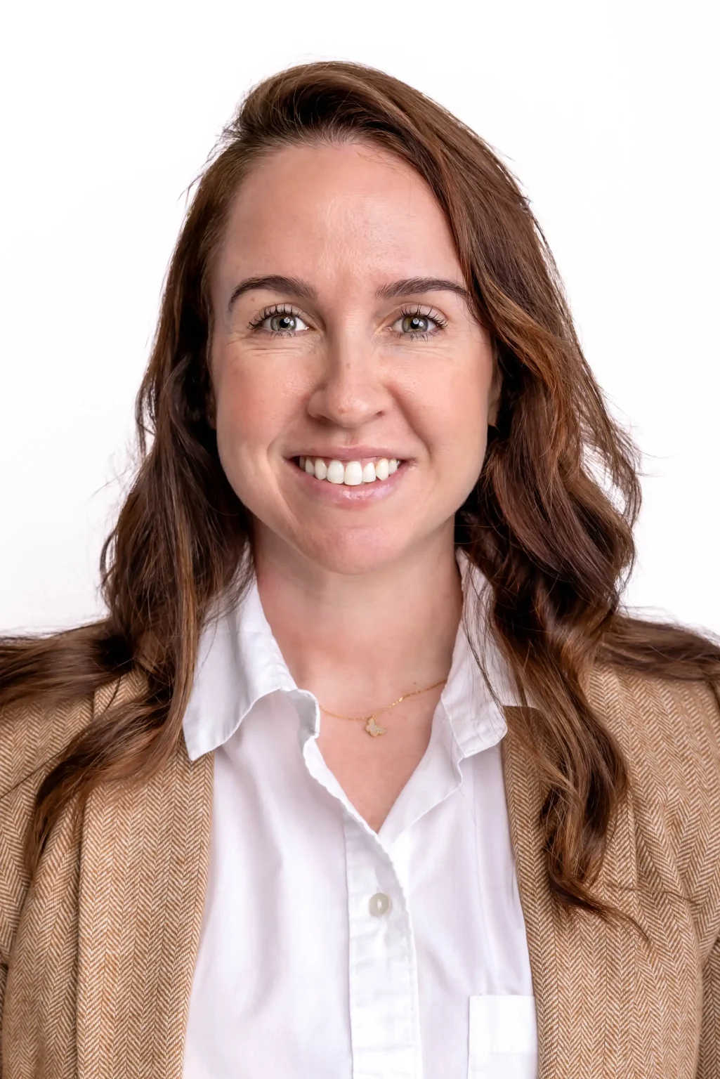 A woman with long brown hair smiles at the camera, wearing a white collared shirt and a tan jacket, against a plain white background.