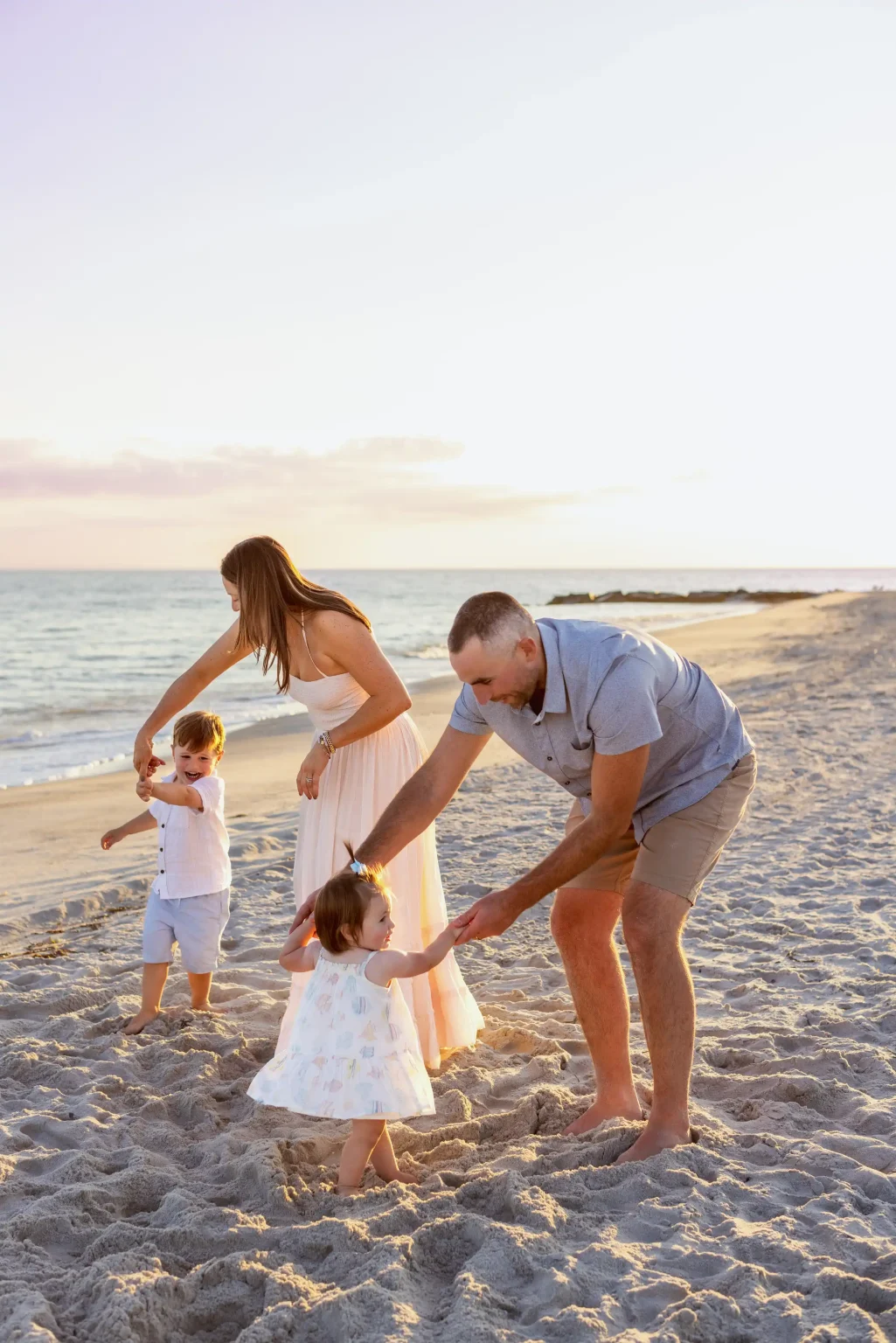 Two adults and two young children walk and play together on a sandy beach near the shoreline during sunset.