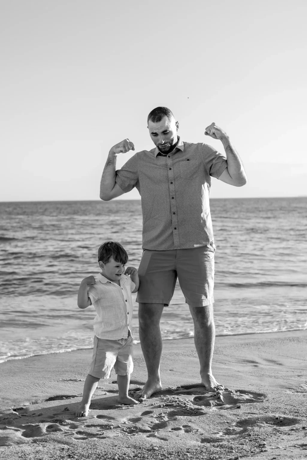 Man and young boy stand on a beach, both flexing their arms and facing forward. The ocean and sky are visible in the background. Image is in black and white.