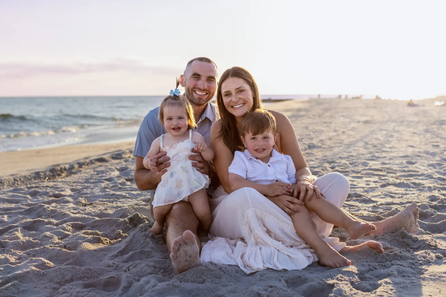 A family of four sits together on a sandy beach near the ocean, smiling at the camera during sunset.