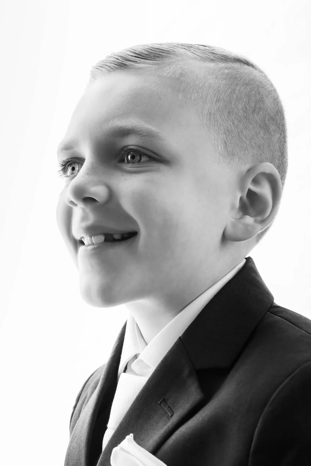 Young boy in a suit and tie, smiling and looking slightly to the left, photographed in black and white against a plain background.