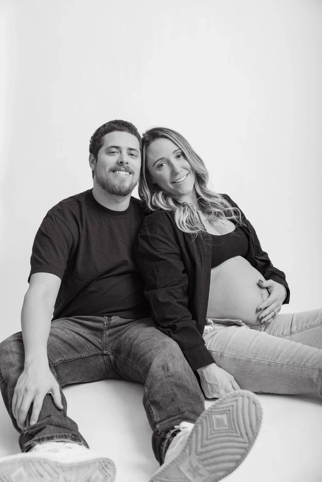 A man and a pregnant woman sit on the floor against a plain backdrop, smiling at the camera. The woman rests her hand on her exposed belly.