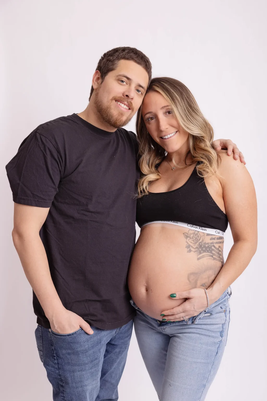 A man and a pregnant woman stand together smiling, with the woman's hand on her bare belly. They are casually dressed against a plain light background.