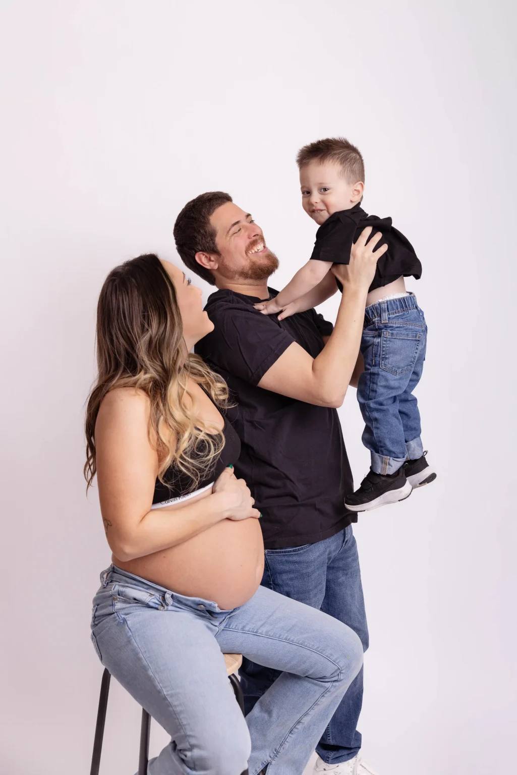 A pregnant woman sits on a stool, while a man holds a smiling young boy in his arms. All three are casually dressed against a plain white background.