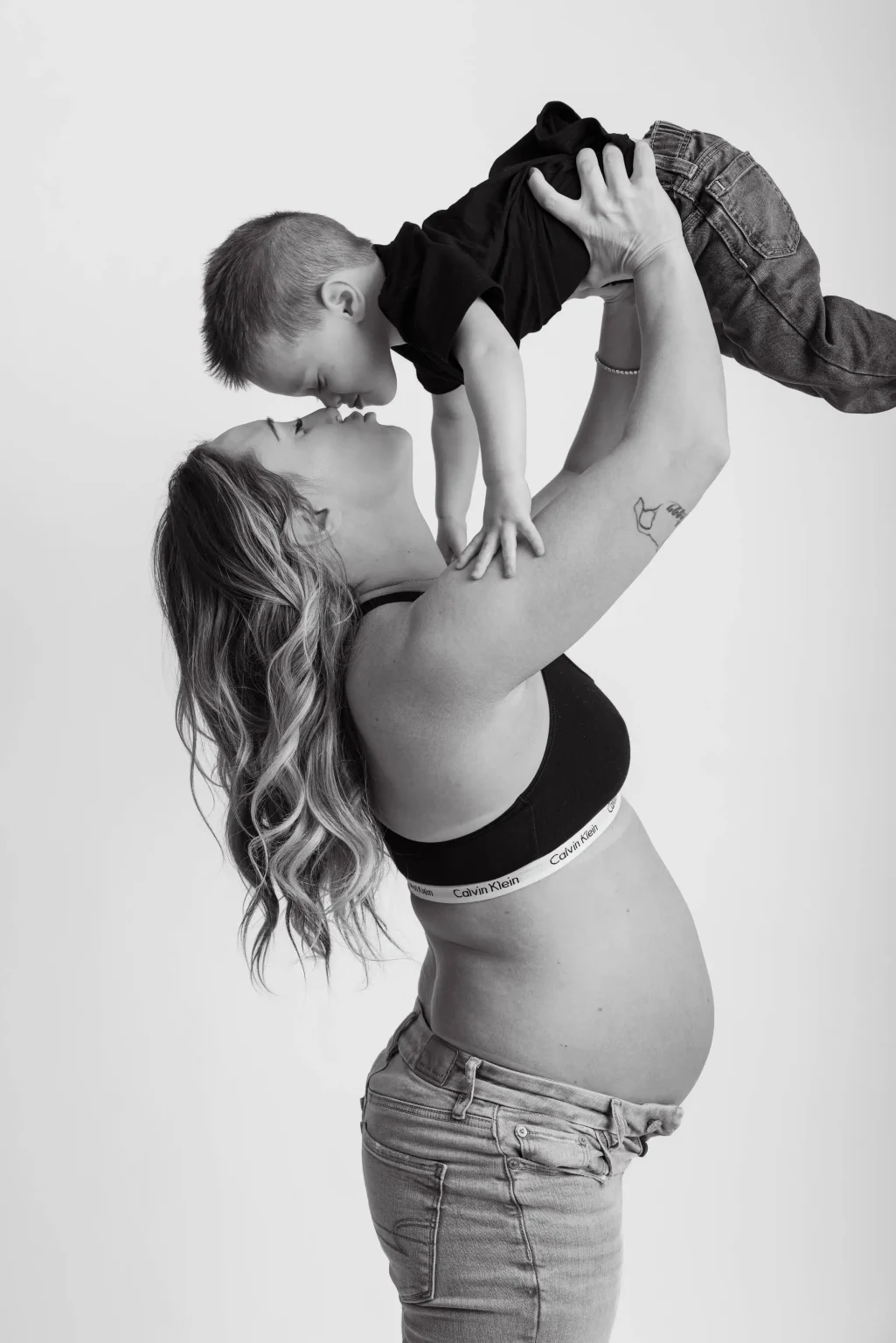 A pregnant woman in a sports bra and jeans lifts a young boy above her, both touching noses. The background is plain white.