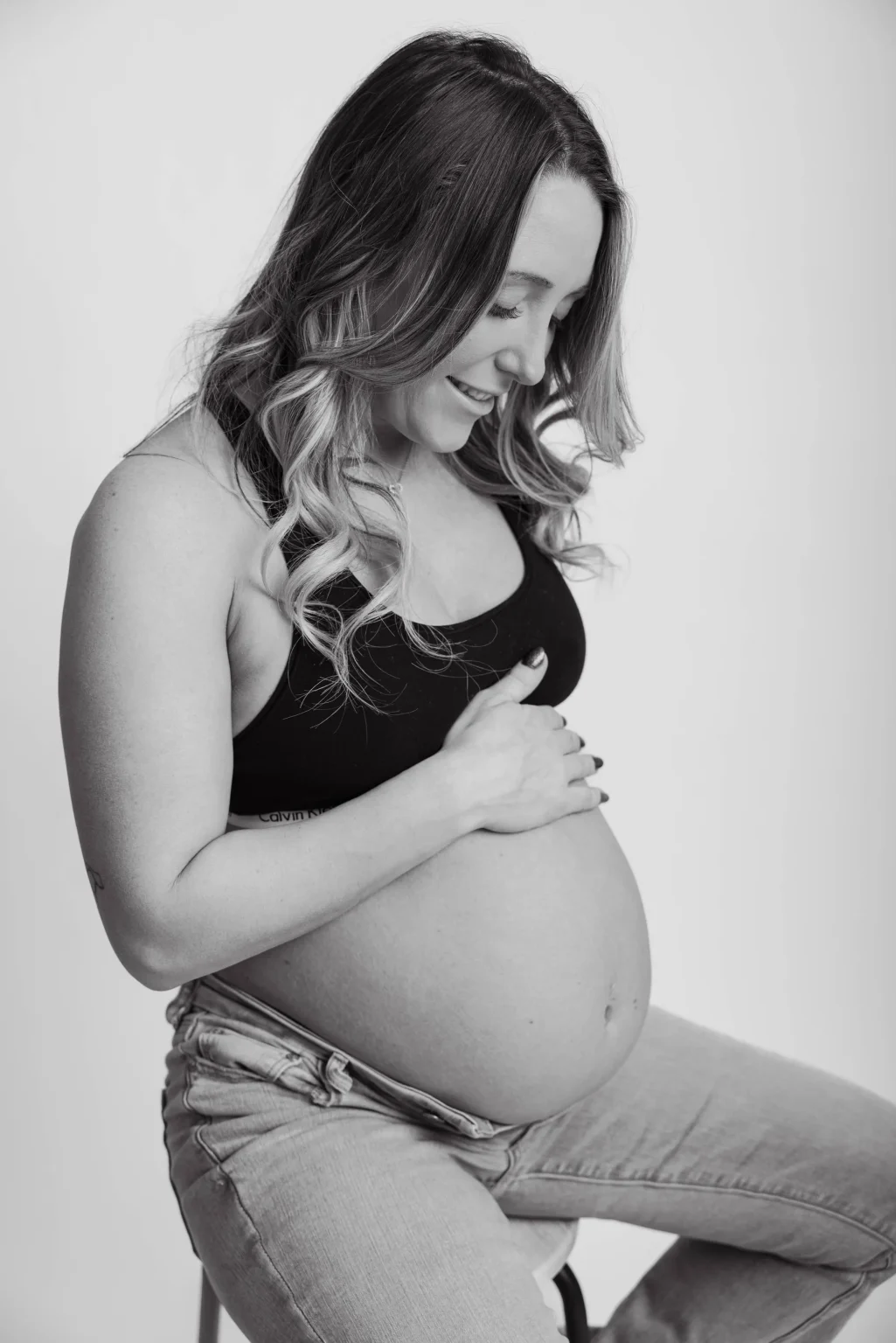 A pregnant woman in a black bra and jeans sits on a stool, touching her bare belly and looking down, photographed in black and white.