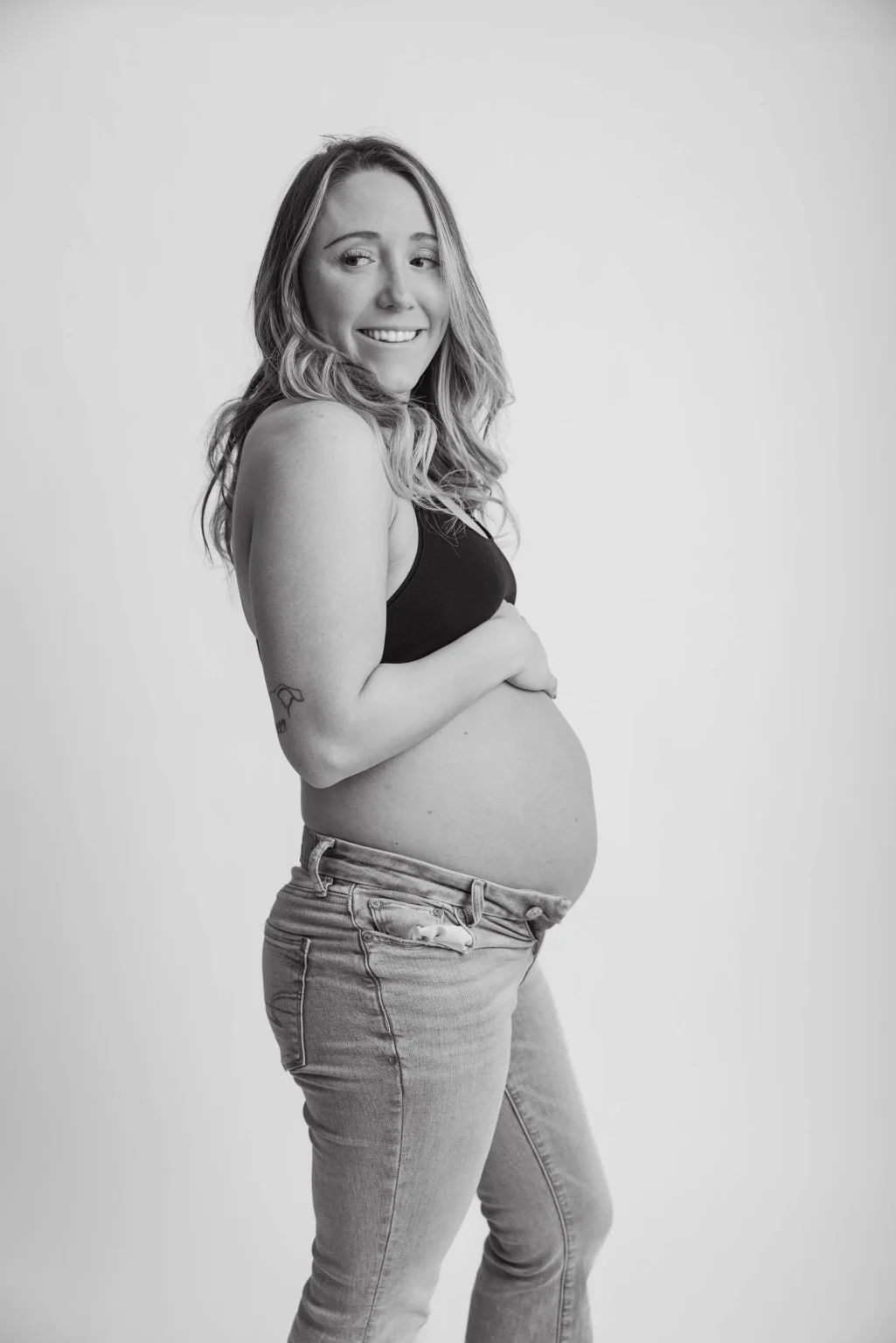 Pregnant woman in a black top and jeans stands sideways, smiling, with one hand on her bare belly against a plain background.