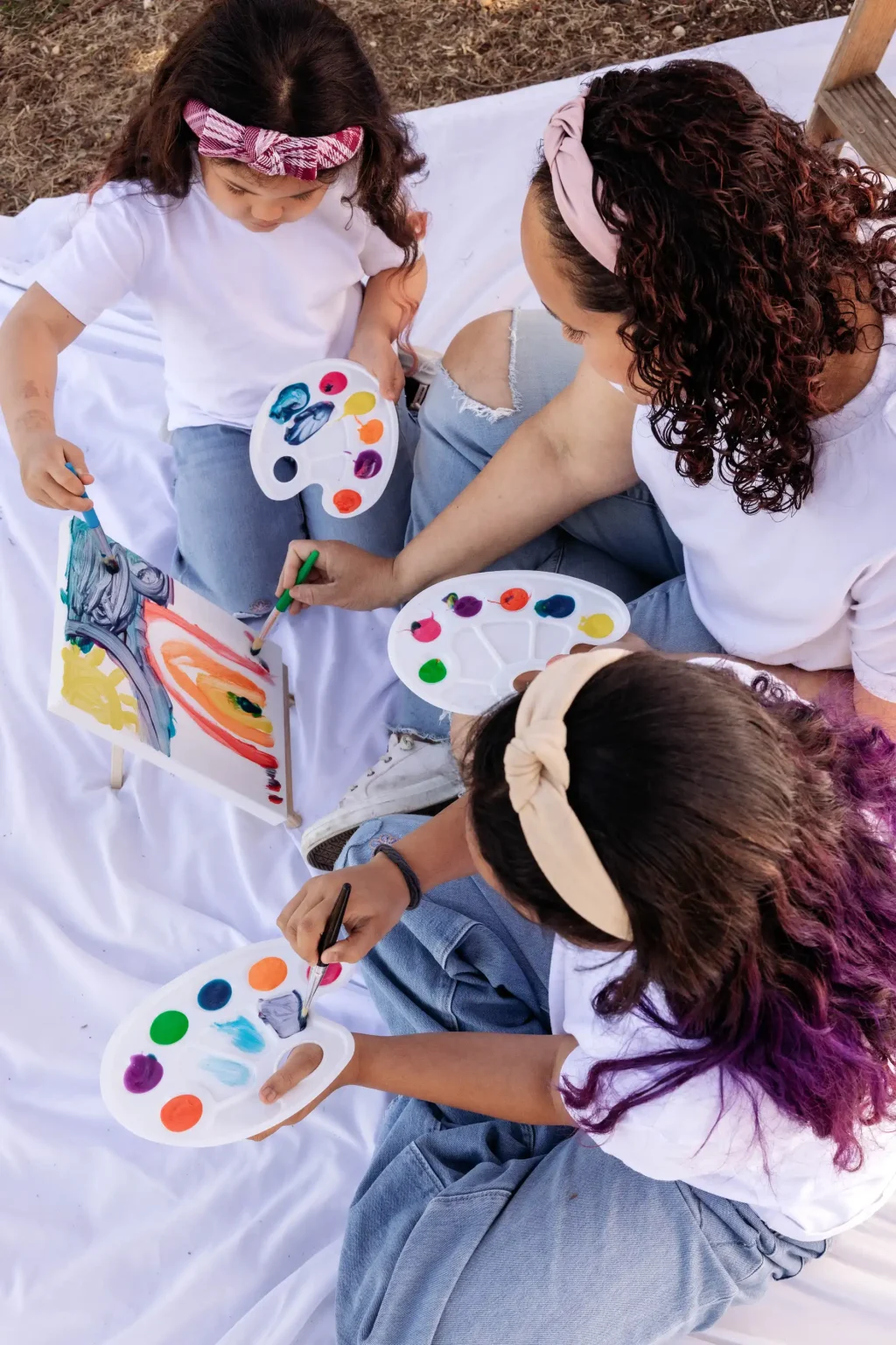 Three people sit on a white blanket outdoors, painting on canvases with palettes of colorful paints.