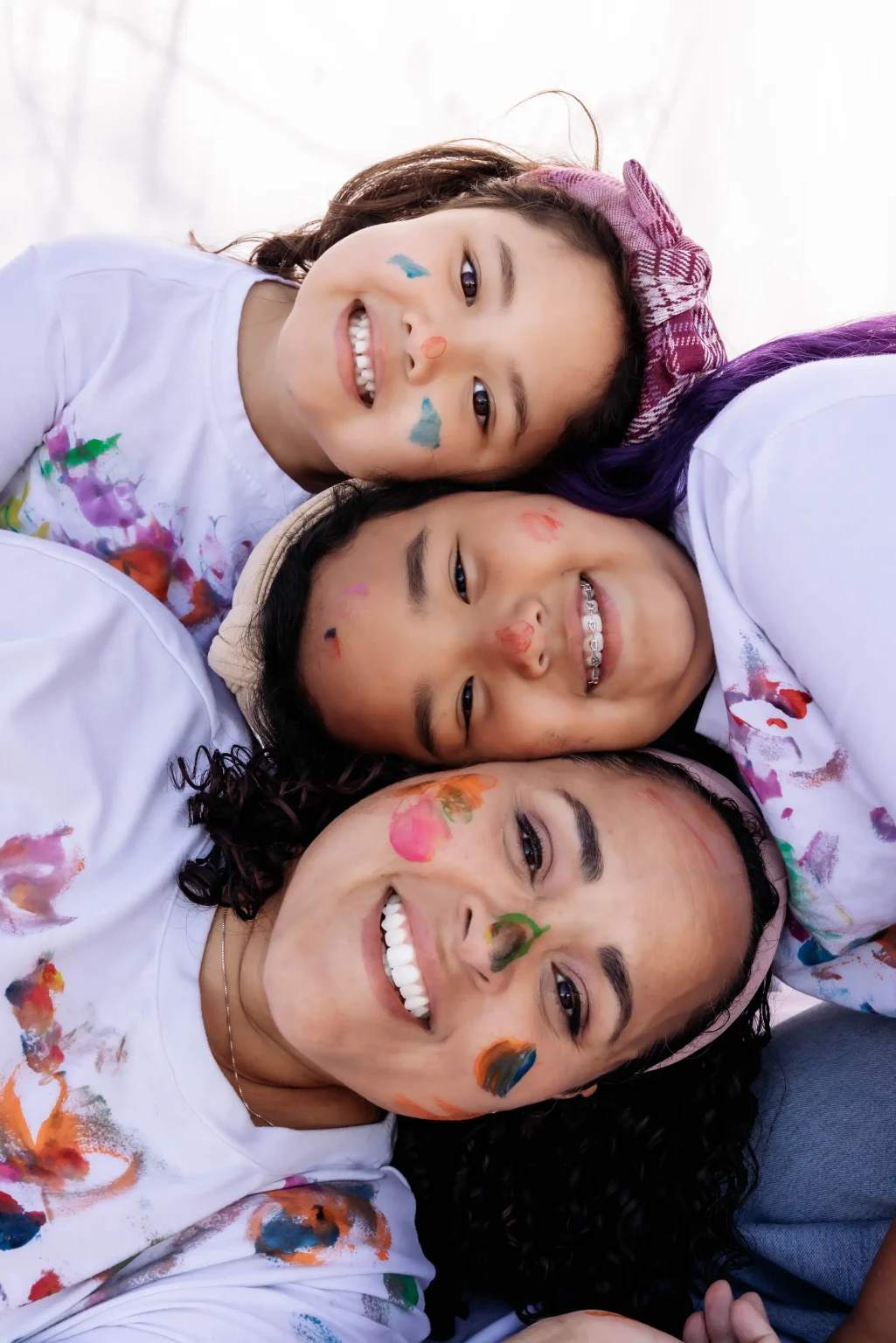 Three people, two children and one adult, lie close together smiling, with colorful paint on their faces and white shirts.