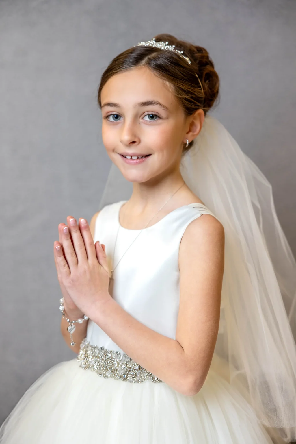 Young girl in a white dress and veil standing with hands pressed together in prayer, wearing a headband, necklace, and bracelet, posing for a formal portrait against a neutral background.