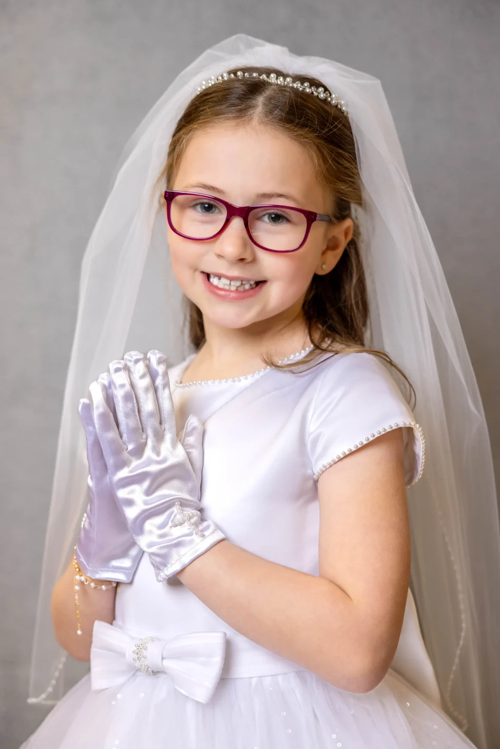 A young girl wearing glasses, a white dress, gloves, and a veil smiles at the camera with her hands together in front of her.