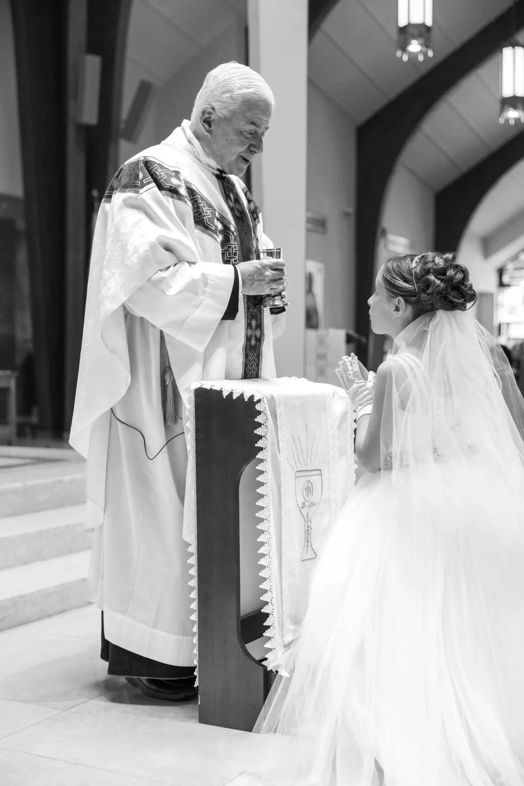 A priest offers communion to a girl in a white dress and veil kneeling at an altar in a church.