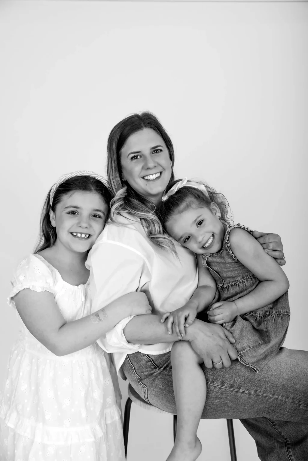 A woman sits smiling with two young girls, one in a light dress standing beside her and the other in a sleeveless dress sitting on her lap, against a plain background.