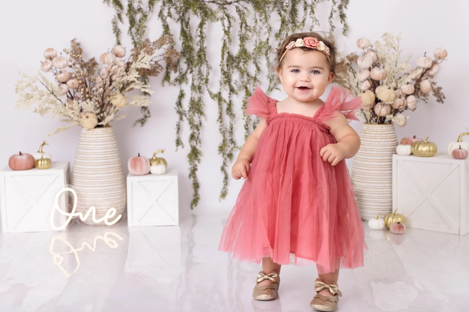 A baby in a pink dress stands in front of autumn-themed decorations with a neon sign that says "One," indicating a first birthday celebration.