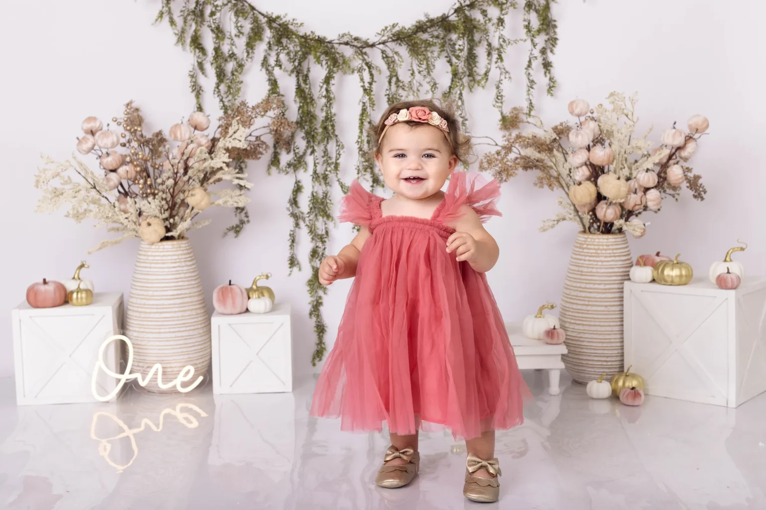 A toddler in a pink tulle dress stands smiling in front of autumn-themed decorations with a sign that says “One” beside her.