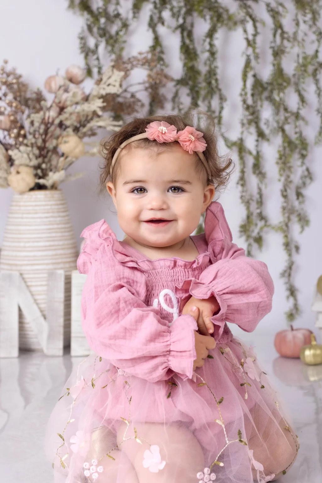 A smiling baby wearing a pink dress and floral headband stands indoors, with a vase of dried flowers and decorative letters in the background.